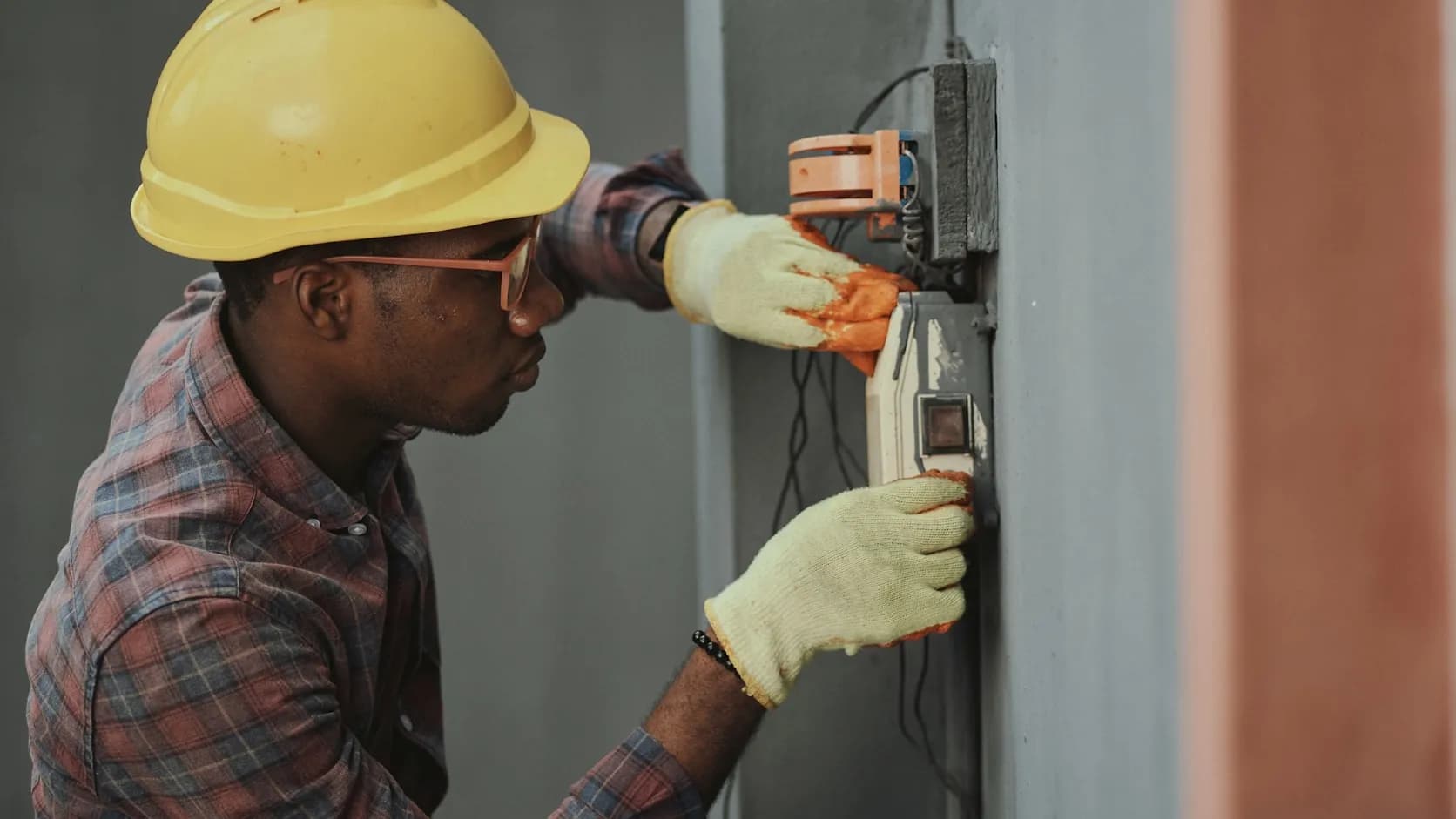 An electrician in a hard hat and gloves at work, both hands occupied wiring a wall-mounted electrical panel.