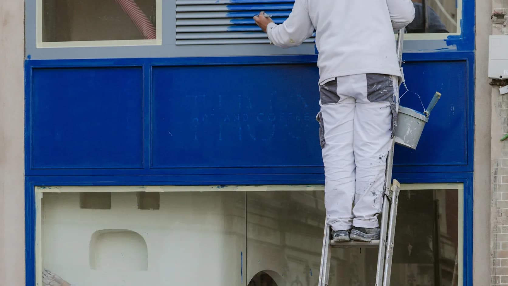 A house painter in white overalls stands on a ladder, brush and paint pail in hand, at work coating a blue exterior wall.