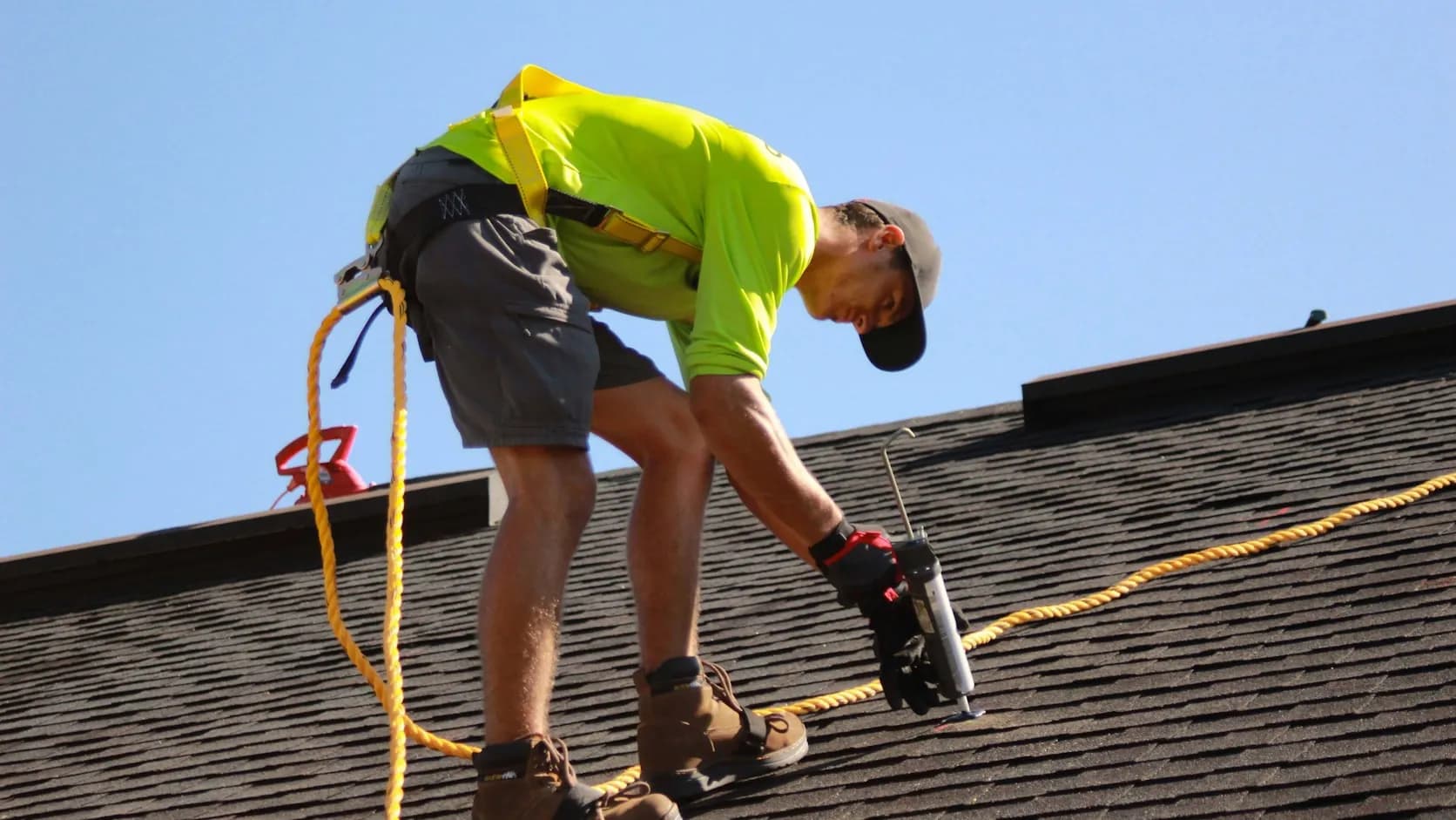 A harnessed roofer kneeling on an asphalt-shingle roof, running a nail gun to fasten shingles — a roofer at work.