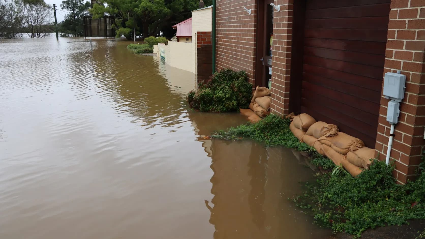 Brown floodwater rising against a brick home with sandbags stacked at the door — a residential water-damage emergency a restoration crew would respond to; no worker is visible in the frame.