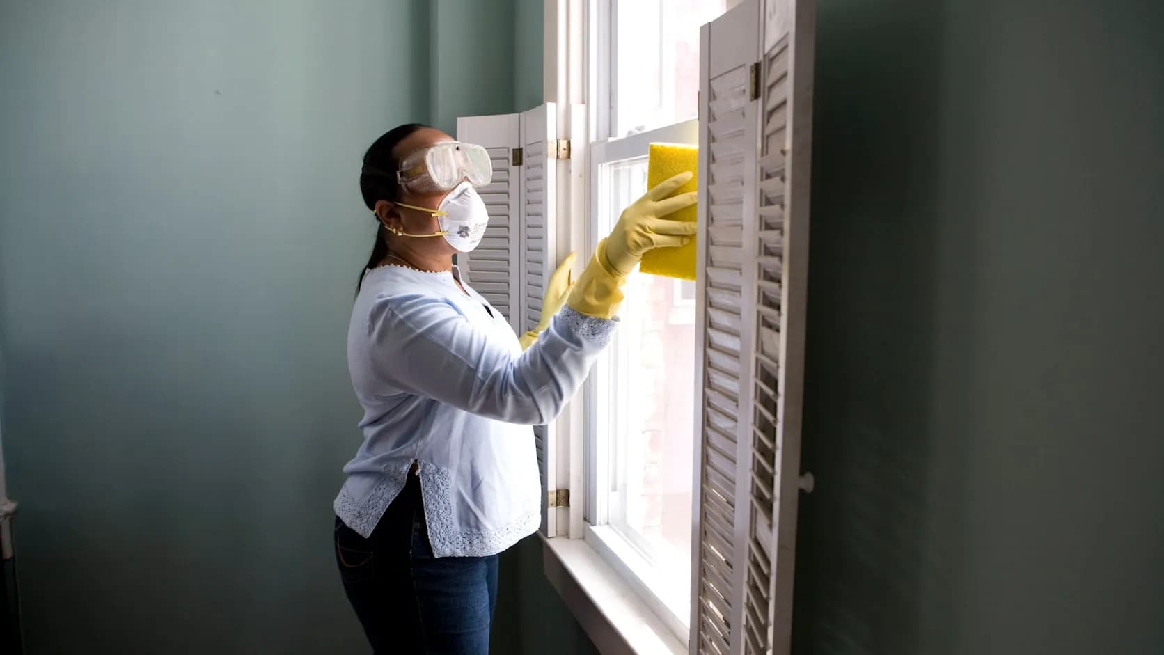 A house cleaner in a mask and rubber gloves at work, scrubbing a window inside a home.