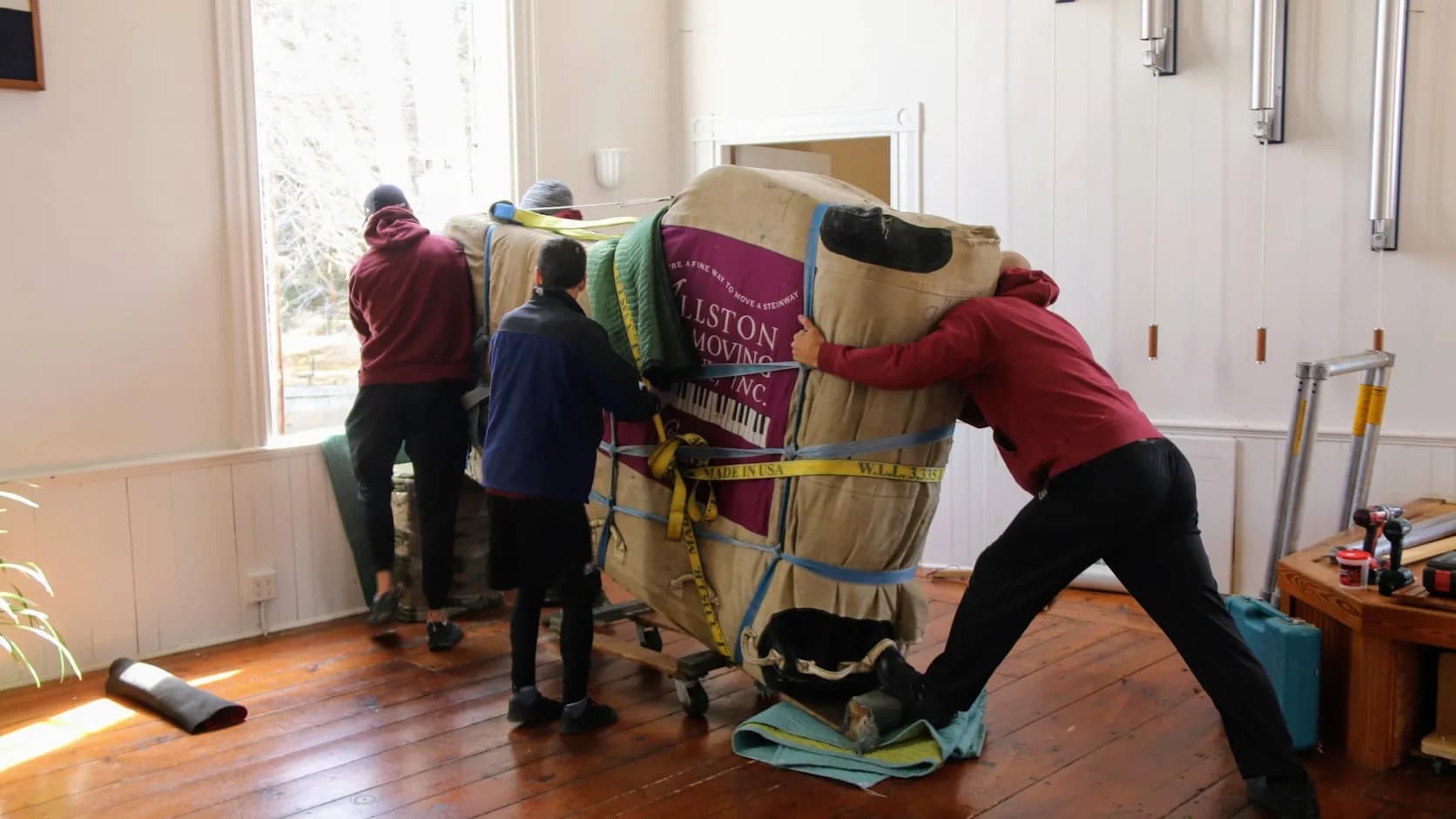 A hauling crew carrying a large wrapped load on a dolly out of a house, workers actively at work (reads as a moving/junk-removal crew moving a heavy item).