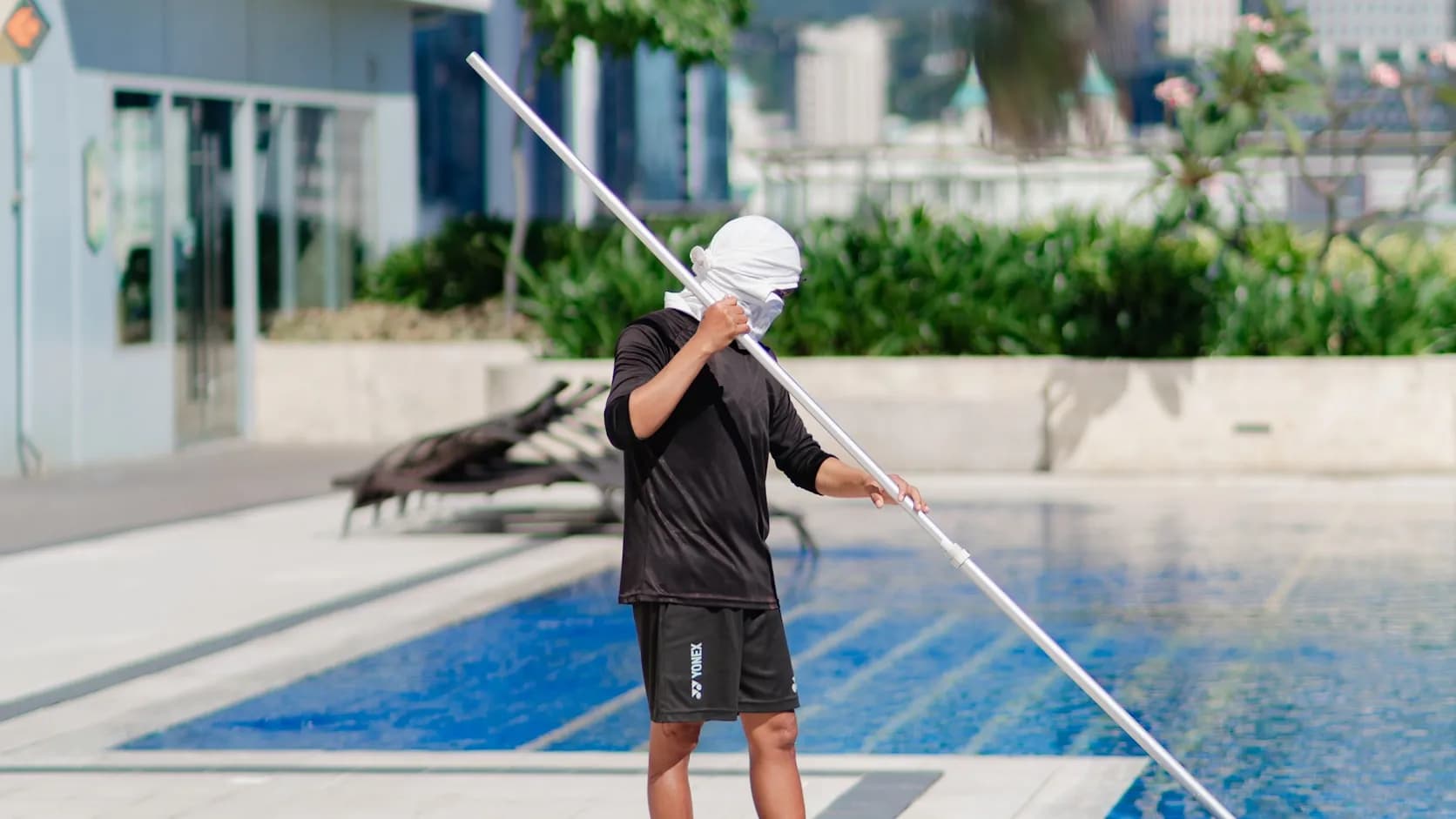 A pool service worker at the poolside gripping a long telescopic cleaning pole, mid-job on a sunny day.